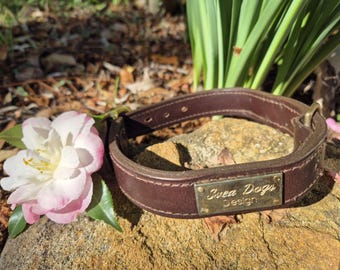 May include: A brown leather dog collar with a brass buckle and a brass nameplate that reads "Svea Dogs Design." A pink and white flower rests beside the collar on a stone.