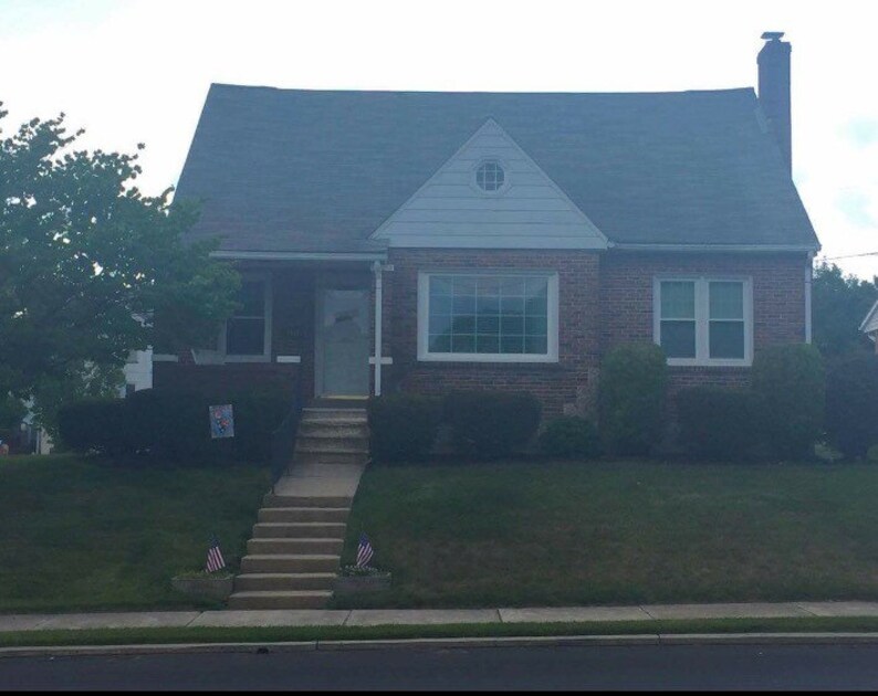 Puede incluir: Una casa de ladrillo con un techo gris y una puerta delantera blanca. La casa tiene dos ventanas en la parte delantera y una peque&ntilde;a ventana en el hastial. Hay un c&eacute;sped verde delante de la casa con un conjunto de escaleras que conducen a la puerta delantera.