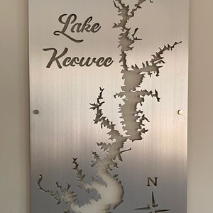 May include: A brushed silver metal sign with a cut-out design of Lake Keowee, a compass rose, and the text "Lake Keowee".