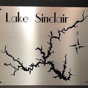 Può includere: Un cartello in metallo spazzolato con una mappa ritagliata dettagliata del lago Sinclair. Il contorno del lago è nero su uno sfondo argentato. Il nome "Lake Sinclair" è ritagliato in alto, con una rosa dei venti e la lettera "N" per il nord.