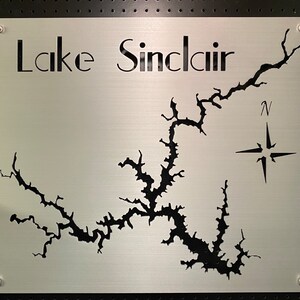 Può includere: Un cartello in metallo argentato con il testo "Lake Sinclair" e un ritaglio nero della forma del lago. Una rosa dei venti si trova nell'angolo in alto a destra.