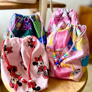 May include: Three drawstring bags with different patterns. The first bag is pink with white polka dots and a red and white Minnie Mouse design. The second bag is pink with a princess design. The third bag is blue and green with a castle design.