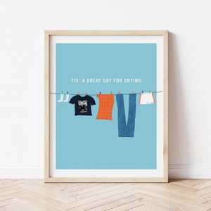Può includere: Un poster azzurro chiaro con il testo "Tis' a great day for drying" e una fila di vestiti appesi a uno stendino. I vestiti includono una maglietta, un paio di jeans, un paio di pantaloncini e due calzini.