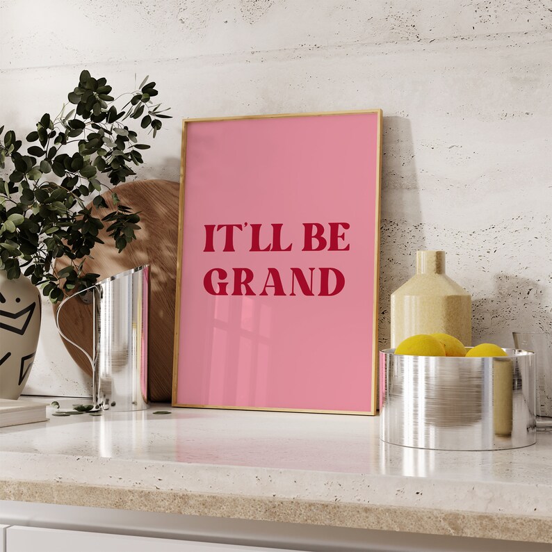 Puede incluir: Una impresi&oacute;n enmarcada en rosa con el texto "IT'LL BE GRAND" en letras rojas. La impresi&oacute;n est&aacute; sobre una encimera blanca con una tabla de cortar de madera y una jarra de plata.
