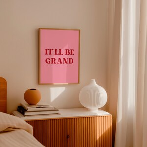 Puede incluir: Una impresi&oacute;n enmarcada rosa con el texto "It'll be grand" en letras rojas, colgada en una pared blanca sobre una mesita de noche de madera con un jarr&oacute;n blanco y libros.