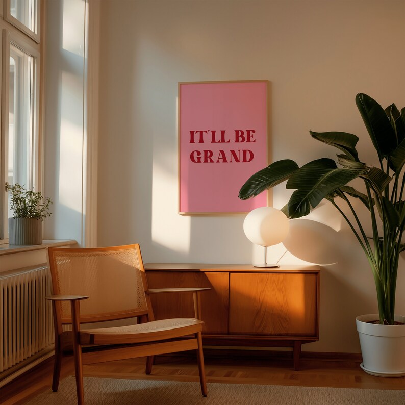 Puede incluir: Un p&oacute;ster enmarcado en rosa con el texto "It'll be grand" en letras rojas, colgado en una pared de una sala de estar. Tambi&eacute;n se puede ver una silla de madera, un armario de madera, una l&aacute;mpara y una planta en maceta en la imagen.