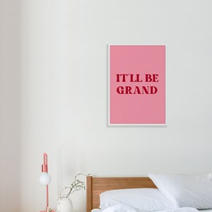 Puede incluir: Una impresi&oacute;n enmarcada rosa con texto blanco que dice "IT'LL BE GRAND" en una pared blanca sobre una cama con ropa de cama blanca y una cabecera de madera.