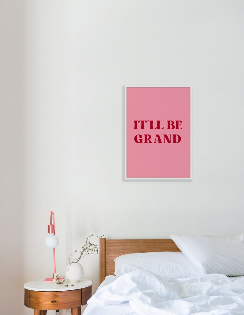 Puede incluir: Una impresi&oacute;n enmarcada rosa con texto blanco que dice "IT'LL BE GRAND" en una pared blanca sobre una cama con ropa de cama blanca y una cabecera de madera.