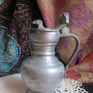 Könnte beinhalten: Ein antiker Krug aus Zinn mit einem Klappdeckel und einem Griff. Der Deckel hat einen dekorativen Widderkopf. Der Krug steht auf einer weißen Spitzendecke. Der Hintergrund umfasst farbenfrohe Textilien.