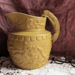 May include: A yellow ceramic pitcher with a detailed embossed design of a hunting scene. The pitcher has a unique handle shaped like a deer's head.