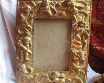 May include: A gold picture frame with a carved design of cherubs and ribbons. The frame is ornate and has a vintage look.