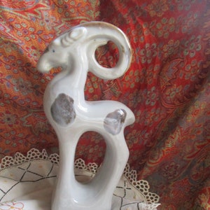 May include: A white ceramic figurine of a goat with brown spots and a curled horn. The figurine is standing on a white crocheted doily with embroidered flowers.