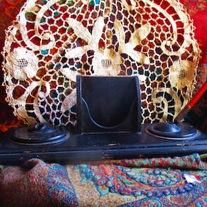 May include: A vintage black inkwell with two ink pots and a pen holder. A red pen is placed in one of the ink pots. A cream lace doily and a colourful patterned textile are in the background.