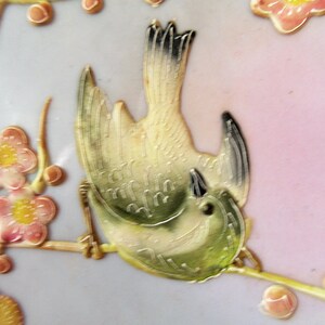 May include: A detailed ceramic bird with green and yellow plumage, perched on a branch with pink blossoms. The bird is depicted in a stylized, almost abstract manner, with its wings spread wide and its head tilted back.