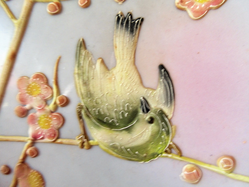 May include: A detailed ceramic bird with green and yellow plumage, perched on a branch with pink blossoms. The bird is depicted in a stylized, almost abstract manner, with its wings spread wide and its head tilted back.