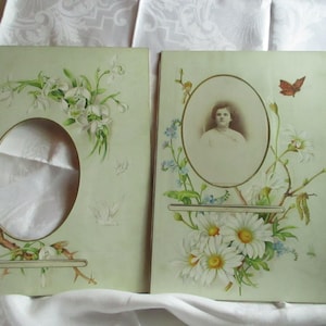 May include: Two antique photo frames with oval openings. One frame features a floral design with white flowers and green leaves. The other frame holds a sepia-toned portrait and is decorated with daisies and a butterfly.