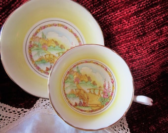 May include: A vintage teacup and saucer set. The cup and saucer are pale yellow with a gold rim. The center of each piece features a colorful landscape scene with a bridge, houses, and flowers. The set is placed on a white lace doily.