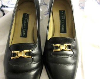 May include: A pair of black leather Naturalizer loafers. Each shoe features a gold-colored buckle and a rounded toe. The interior of each shoe has a tan insole with the Naturalizer brand name.