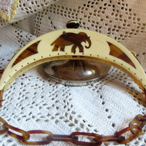 May include: A vintage, cream-colored, half-moon shaped purse frame with a brown elephant design. The frame is attached to a brown chain.