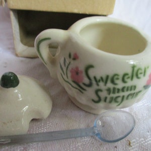 Può includere: Zuccheriera in ceramica bianca con un design floreale verde e rosa e il testo "Sweeter than Sugar". La zuccheriera ha un coperchio abbinato e un piccolo cucchiaio blu.