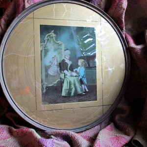 May include: A vintage framed picture with a gold-coloured frame and a scene of a woman in a blue dress reading to a young girl in a white dress. The picture is on a pink and purple patterned fabric.