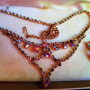 May include: A delicate gold necklace with a cascading design of iridescent pink and clear rhinestones. Matching earrings with a single pink rhinestone dangle.