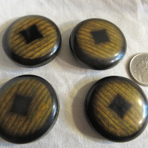 Puede incluir: Cuatro botones redondos, negros y dorados con un diseño cuadrado negro en el centro. Los botones tienen un patrón de veta de madera.