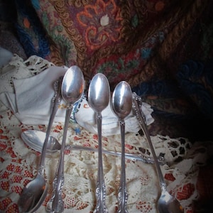 Può includere: Cinque cucchiai d'argento con manici decorati sono disposti su una tovaglia di pizzo bianco con un tessuto a motivi rossi e dorati sullo sfondo.