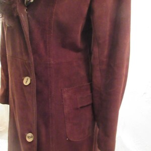 May include: A long, brown suede coat with a fur collar and a single pocket on the right side. The coat has a button closure.