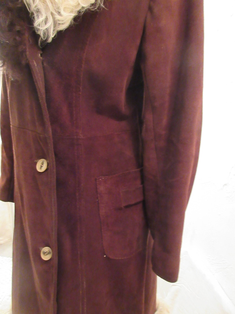 May include: A long, brown suede coat with a fur collar and a single pocket on the right side. The coat has a button closure.