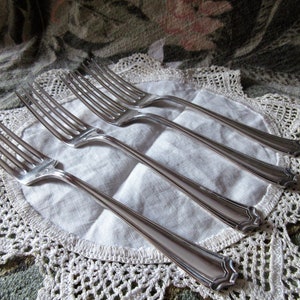 May include: Four silver forks with ornate handles are arranged on a white doily with a lace border.