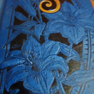 May include: A blue and gold book cover with a floral design. The cover features a large, stylized letter 'S' in gold and the word 'SUN' in gold. The floral design is made up of blue lilies with black outlines. The background is a dark blue color.