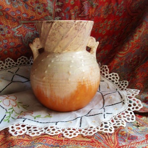 May include: A white and brown ceramic vase with two handles and a textured surface. The vase is sitting on a white lace doily.