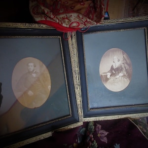 May include: Two antique oval portraits in ornate black and gold frames. The portraits are of a man and a woman, both in formal attire, and are displayed on a dark surface.