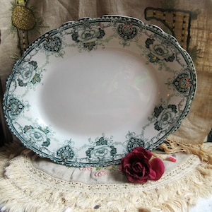 May include: A white porcelain serving platter with a green and black floral design. The platter has a scalloped edge and is decorated with a repeating pattern of flowers and leaves. The platter is sitting on a white lace tablecloth with a fringe.