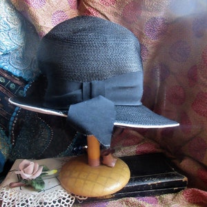 Pode incluir: Um chapéu de palha preto com uma aba larga e uma fita de cetim preto. O chapéu está sentado em um suporte de chapéu de madeira.