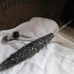 Pode incluir: Um alfinete longo, fino e preto com uma conta preta na ponta. O alfinete está coberto de pequenas contas prateadas. O alfinete está sobre uma superfície de tecido branco.