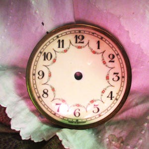 May include: A white clock face with black Roman numeral hour markers and floral accents. The clock face is surrounded by a gold-coloured metal frame. The clock face reads "Made in Germany".