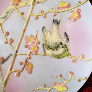 May include: A white ceramic plate with a delicate floral design featuring pink and yellow flowers and a small bird. The plate has a light blue background and a subtle pink rim.
