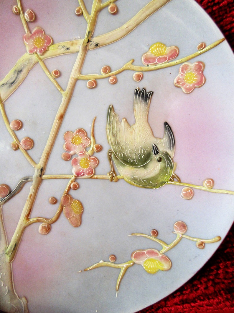 May include: A white ceramic plate with a delicate floral design featuring pink and yellow flowers and a small bird. The plate has a light blue background and a subtle pink rim.