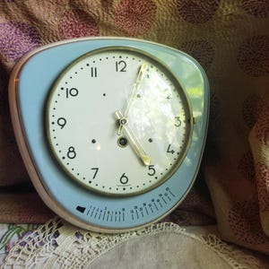 May include: A vintage, light blue and white clock with a rounded, triangular shape. The clock face has black numbers and hands, with a gold-colored trim. A timer dial is at the bottom. The clock is set against a patterned fabric background.