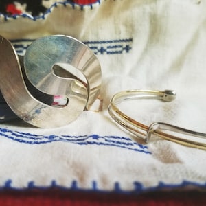 May include: A silver brooch with a swirling design and a two-tone bracelet are displayed on a white cloth with blue embroidery. The brooch has a curved shape, and the bracelet has silver and gold bands. The items are on a vintage-style background.