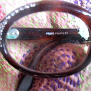 May include: Close-up of a pair of brown tortoiseshell eyeglasses with a rhinestones detail on the frame. The glasses have a black plastic frame and the text "FRAME FRANCE KZ" is visible on the inside of the frame.