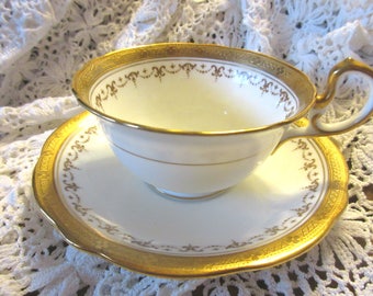 Puede incluir: Una taza de té y un platillo blancos con un borde dorado y un intrincado patrón floral dorado. La taza y el platillo están sobre un mantel de encaje blanco.
