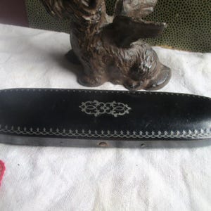 May include: Black oval box with white stitching and a decorative design. A small, brown cast iron dog statue is in the background. The box is on a white cloth.
