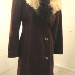 May include: A long, brown suede coat with a large, fluffy white and black fur collar. The coat has a double-breasted closure with gold buttons and a fur trim at the bottom.