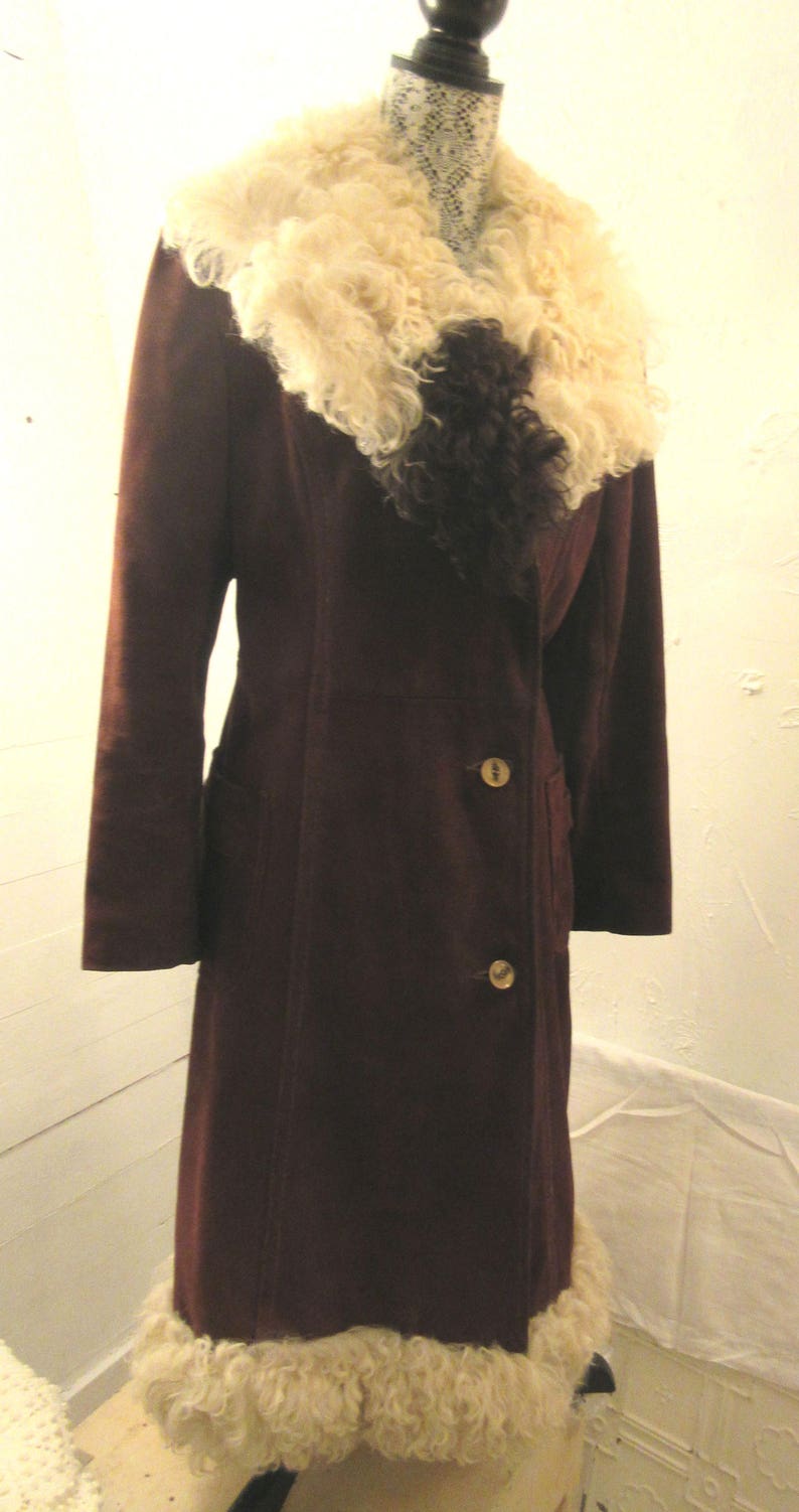 May include: A long, brown suede coat with a large, fluffy white and black fur collar. The coat has a double-breasted closure with gold buttons and a fur trim at the bottom.