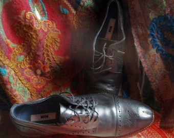 May include: Two pairs of black leather lace-up shoes with brogue detailing. The shoes are sitting on a colorful patterned fabric.