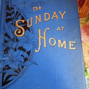 May include: The Sunday at Home book cover with a blue background and gold lettering. The cover features a decorative design of black lilies and leaves in the upper left corner.