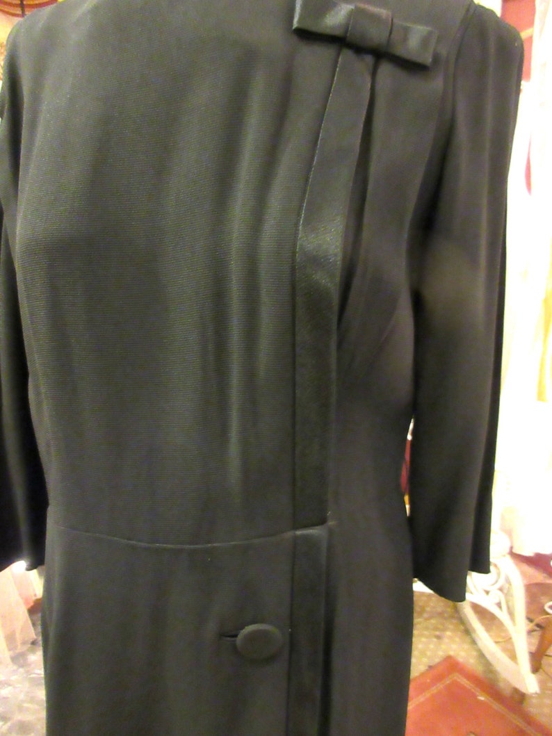 May include: A black dress with a satin trim and a bow at the neckline. The dress has a single button at the waist.